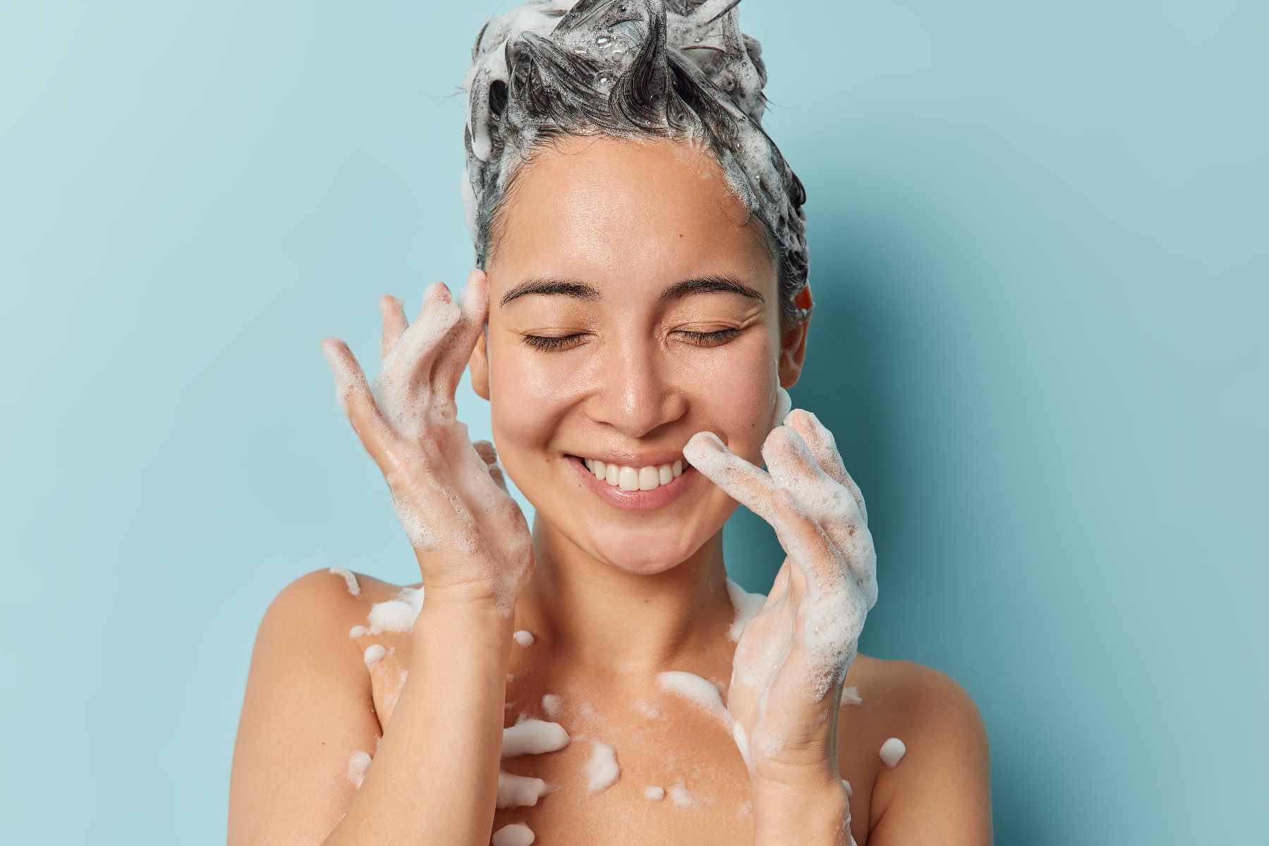 Lachende jonge vrouw met haar ogen dicht met shampoo in haar haren en schuim aan haar handen met een lichtblauwe achtergrond